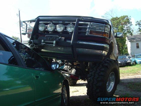 Lifted black truck with a winch and light bar on its front bumper, straddling a green car