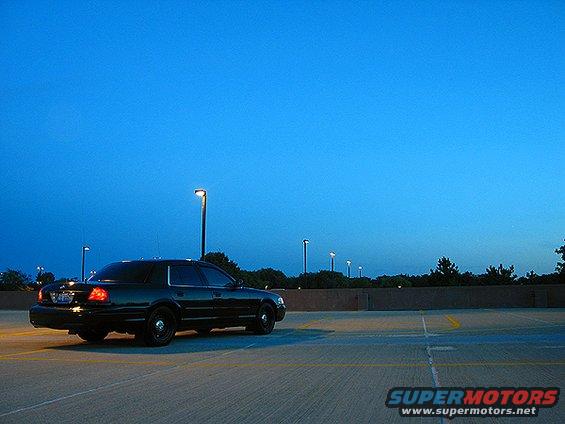Black sedan parked in a lot at dusk. Lights are on in the distance