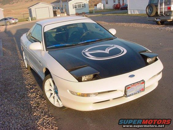 1994 Ford Probe with black hood a large white Mazda logo, pop-up headlights, and aftermarket white wheels