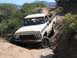 1979 Ford Bronco - A two-tone Ford truck navigates a steep, dusty off-road path with brush and mountains in the background.