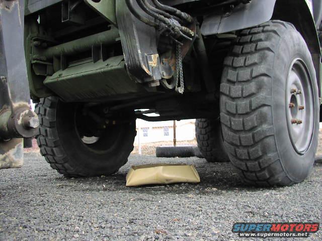 Underside of a military truck showing large tires, suspension, and drivetrain components. A tan object is on the ground beneath the vehicle