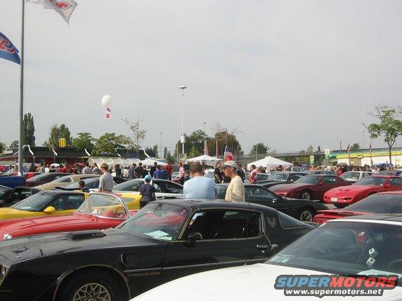 Cars parked at an outdoor event, people milling around. Several classic cars are visible in the foreground and background