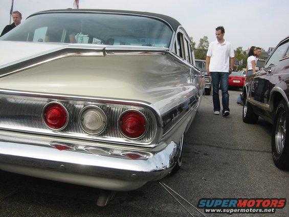 Rear view of a 1959 Chevrolet sedan with triple tail lights and chrome bumper. Another car is parked to the right