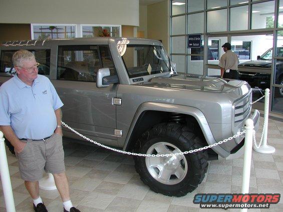 Man stands next to a silver concept SUV with a prominent grille and large tires inside a showroom