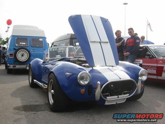 Blue AC Cobra replica with hood open, parked next to a blue van and a red car