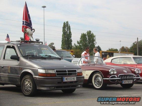 Gray minivan with Confederate flag and bull skull on roof, parked next to a red classic convertible and a red classic coupe