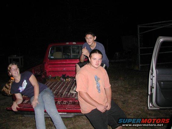 Three people sit in the bed of a red pickup truck at night. A silver SUV door is open to the right
