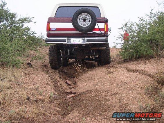 Maroon and white Ford Bronco with a spare tire on the back, driving on a dirt trail