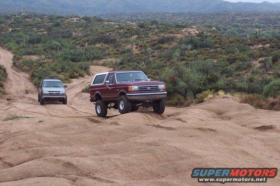 Maroon 1992 Ford Bronco and silver Chevrolet Trailblazer driving on a dirt trail in a desert landscape