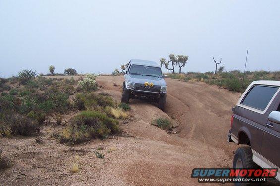 Gray SUV climbing a dirt hill, brown SUV behind it. Desert landscape