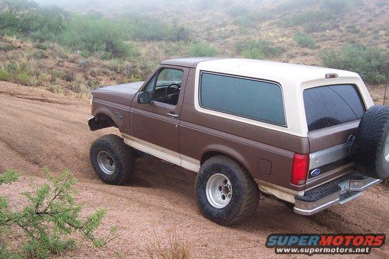 Brown and white Ford Bronco with large tires driving on a dirt trail