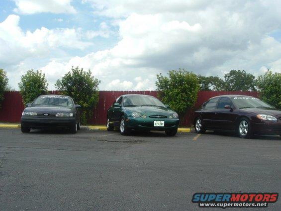 Three Ford Taurus sedans parked in a lot under a cloudy sky. The middle car is green, the left is dark, and the right is maroon