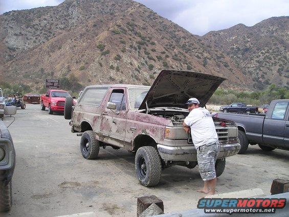 Mud-splattered maroon Ford Bronco with hood open, man leaning on front fender