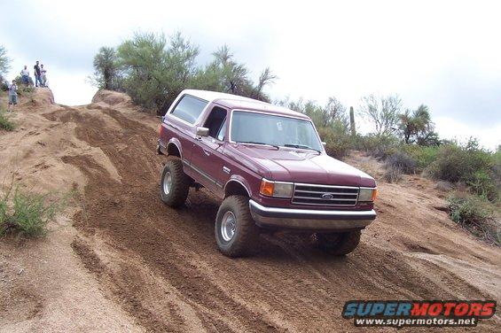 Maroon 1992 Ford Bronco climbing a steep dirt hill with people watching on top
