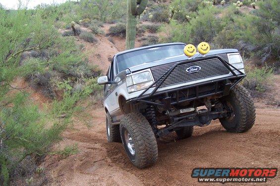 Silver and gray 1992 Ford Bronco with custom bumper and smiley face lights driving on a dirt road