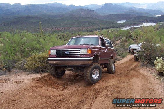 Maroon Ford Bronco with lifted suspension driving on a dirt road in a desert landscape