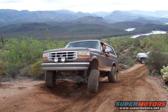 Brown Ford Bronco driving on a dirt trail with a man inside, another SUV behind it, desert mountains in background
