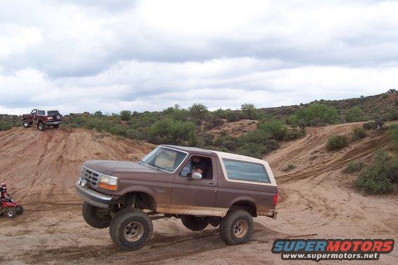 Brown 1992 Ford Bronco with lifted suspension driving on a dirt trail, another SUV on a hill in background