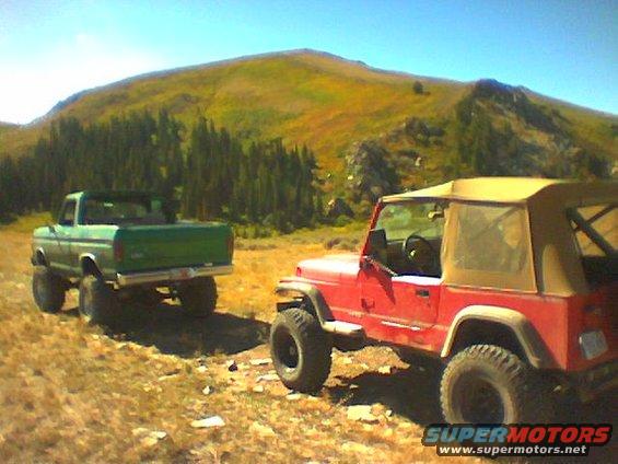 Green 1970s Ford pickup truck and red Jeep Wrangler parked on a dirt road with a mountain in the background