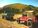 1979 Ford Bronco - Two off-road vehicles, a green Ford pickup and a red Jeep, are parked on a dirt track with a mountain landscape behind them.