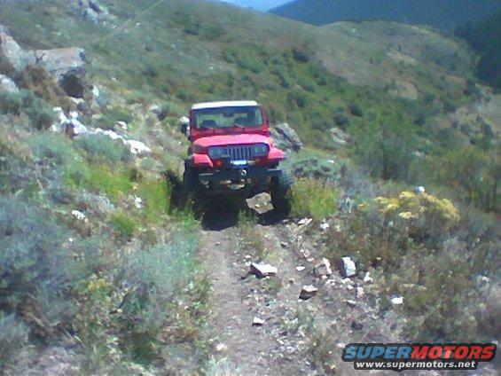 Red Jeep Wrangler driving on a rocky dirt trail in a hilly, vegetated area