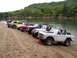 1967 Ford Bronco - Several off-road vehicles are lined up on a sandy shore next to a wide river.