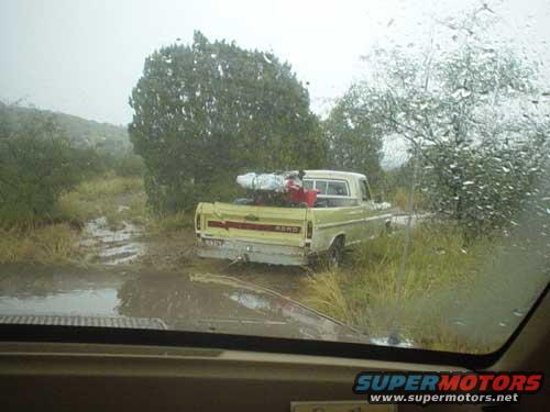 Yellow Ford pickup truck with red and white load in bed, stuck in mud. Taken through a rain-streaked windshield