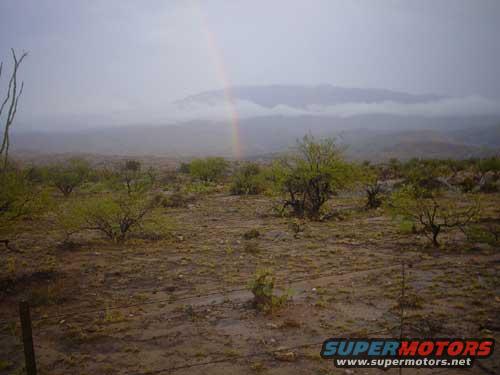 Desert landscape with scrub brush, mountains in background, and a rainbow in the sky. Supermotors.net logo visible