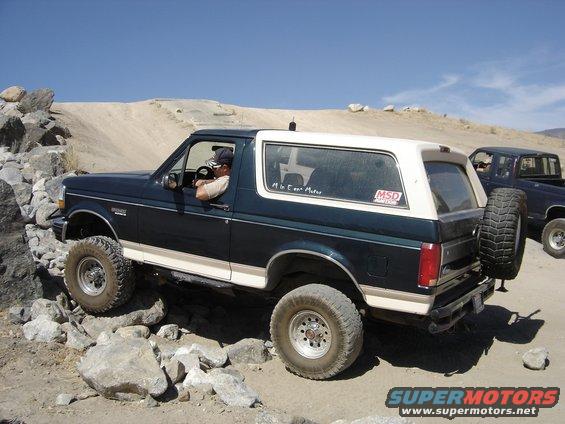 Dark blue and white Ford Bronco navigating rocky terrain. Another dark pickup truck is visible behind it