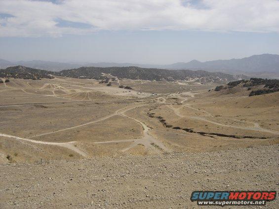 Dirt roads wind through a dry, hilly landscape under a hazy sky; two vehicles are visible in the distance