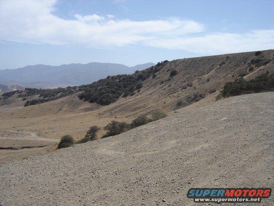 Dry, hilly landscape with scattered trees under a blue sky with clouds. Supermotors logo in lower right