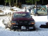 Maroon Ford Bronco stuck in snow, front view. Another green Bronco is visible behind it