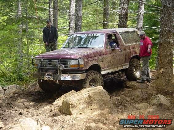 Muddy maroon Ford Bronco navigating an off-road trail with two people nearby