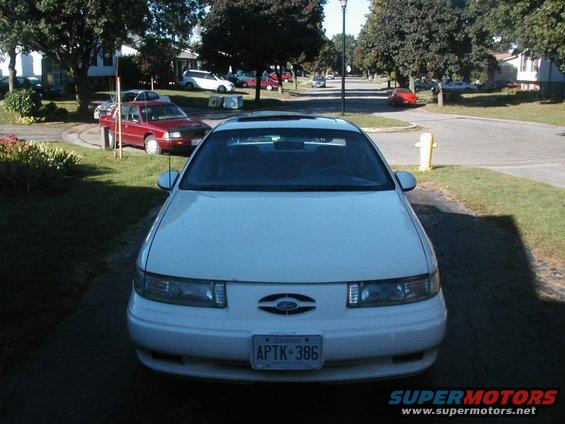 Front view of a white 1993 Ford Taurus parked in a driveway. Another red car is visible in the background