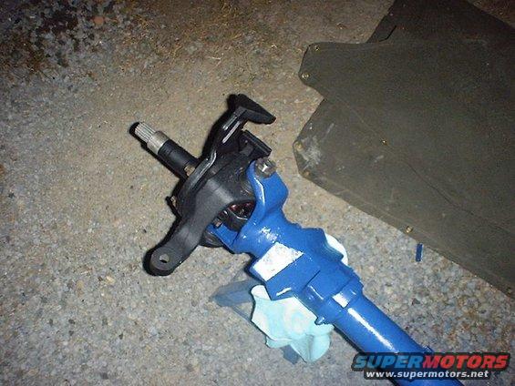 Blue painted steering component with a splined shaft and black bracketry, resting on gravel next to a dark green panel