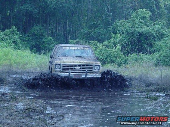 Tan pickup truck with "DIRTY DEEDS OFFROAD" on windshield driving through mud puddle