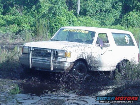 White Ford Bronco with 'DIRTY DEEDS OFFROAD' sticker on windshield stuck in mud