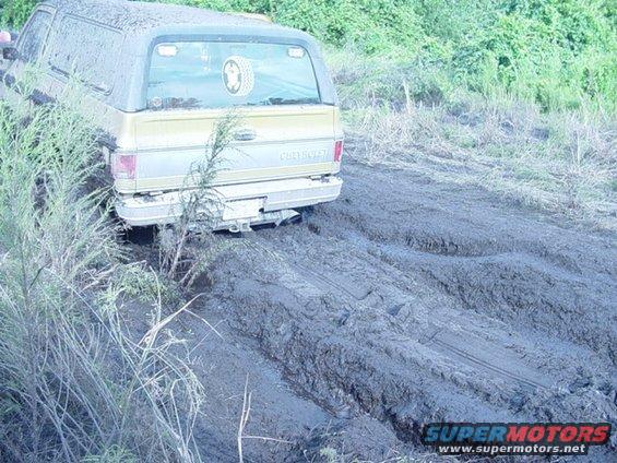 Rear view of a gold Chevrolet K5 Blazer stuck in deep mud, with vegetation around