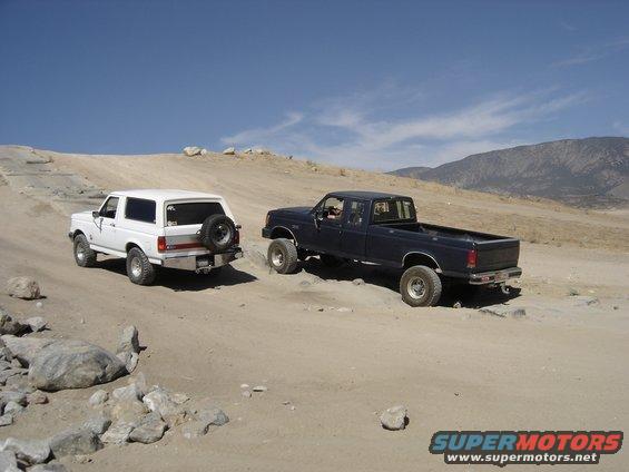 Dark blue pickup truck and white SUV with spare tire on back, driving on dirt road with mountains in background