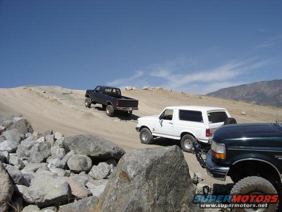 Three off-road trucks, including a dark blue Ford pickup and a white Ford Bronco, navigate a rocky, dusty trail under a clear blue sky