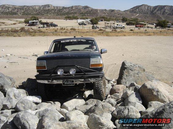 Ford Bronco navigating rocky terrain. Has 'Main Event Motors Las Vegas' on windshield banner and 'Team Trust' sticker on bumper