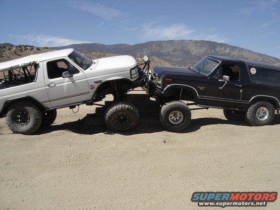 Two lifted Ford Broncos, one white and one dark, are positioned nose-to-nose on a dirt track with mountains in the background
