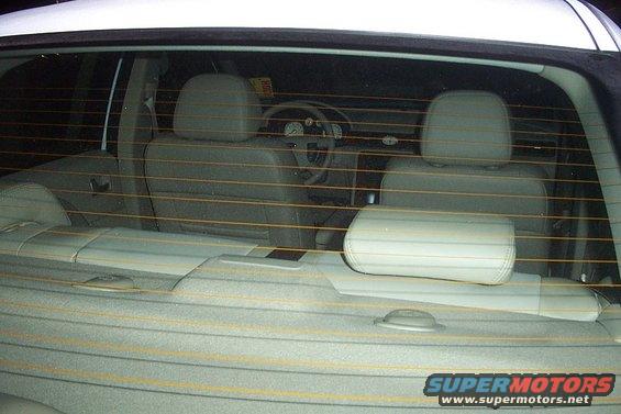 Interior of a white car, seen through the rear window. Light gray seats and headrests are visible, along with the dashboard and steering wheel. A "SUPERMOTORS.NET" watermark is in the lower right
