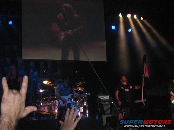 Band playing on stage with a large screen showing a guitarist. Audience members in foreground making rock on