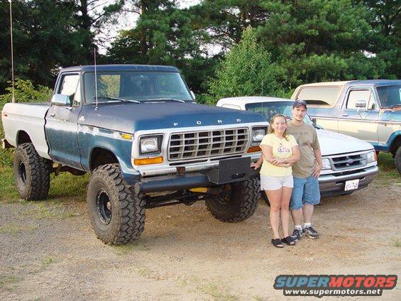 Lifted blue and white Ford F-Series pickup truck with a man and woman standing beside it. Another Ford truck is visible behind