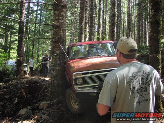 Red pickup truck stuck between trees on a dirt trail, another vehicle visible in background