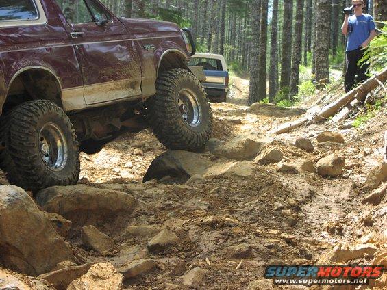 Maroon 1993 Ford Bronco climbing a rocky trail, another Bronco behind it, person filming in background