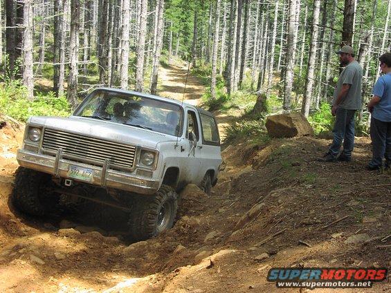 White SUV with large tires navigating a steep, rocky dirt path in a forest. Two people watch from the side