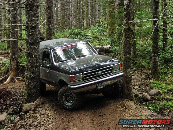 Ford Bronco driving on a dirt trail in a forest, with trees on both sides
