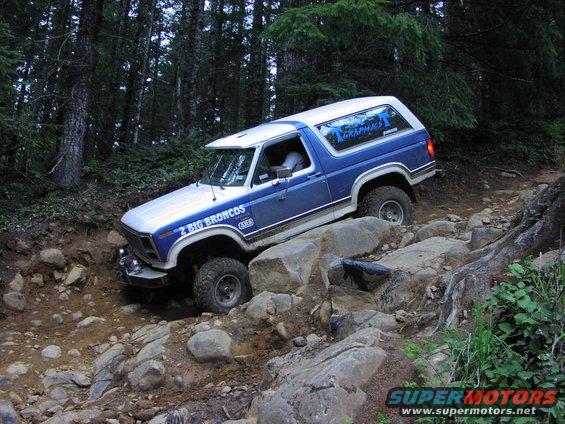 Blue and white Ford Bronco navigating a rocky forest trail, with "2 BIG BRONCOS" text on the side